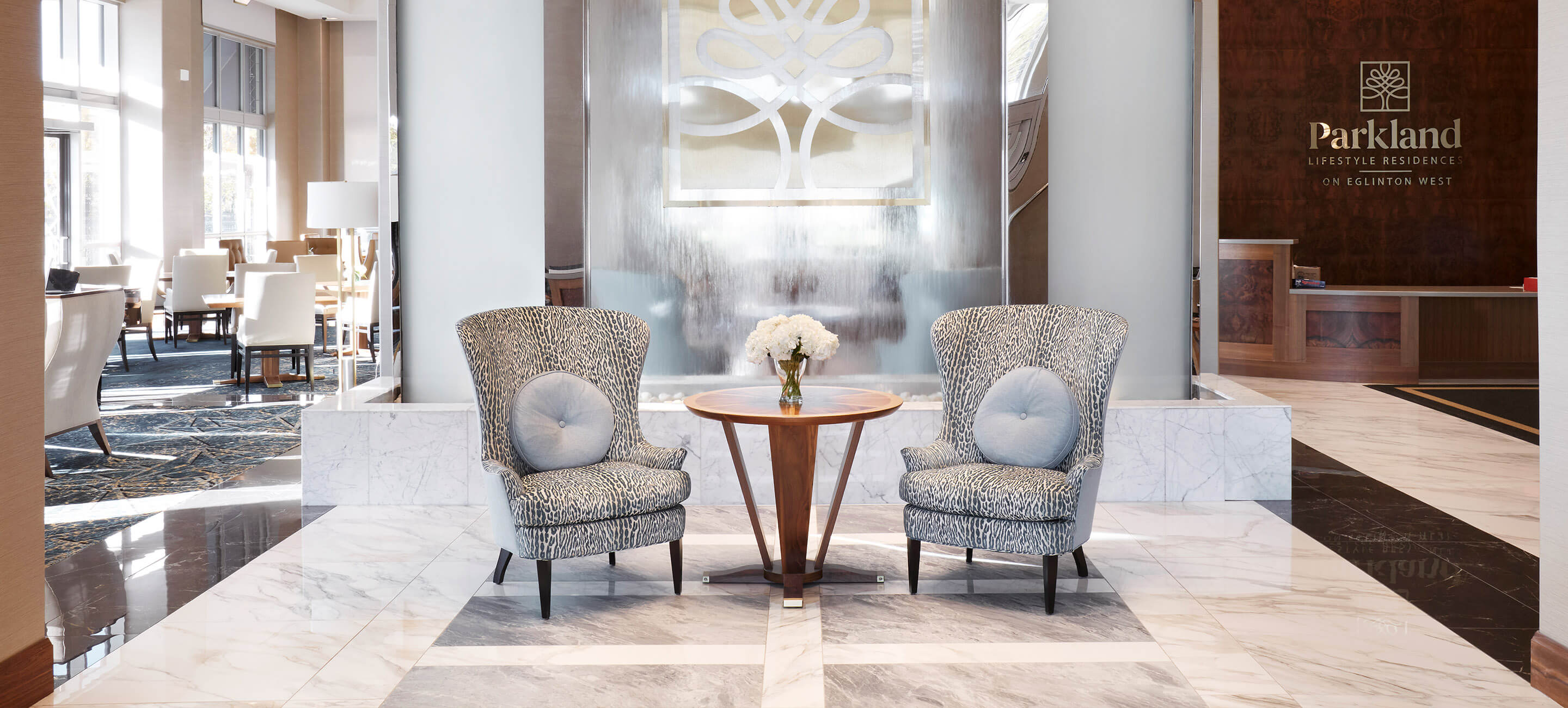Large granite lobby of retirement home. Two chairs with pillows and small table between them are in the center of the image. 